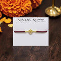 SILVAAS gold-toned Ganesh Ji Rakhi with a maroon thread on a display card, featuring intricate detailing.