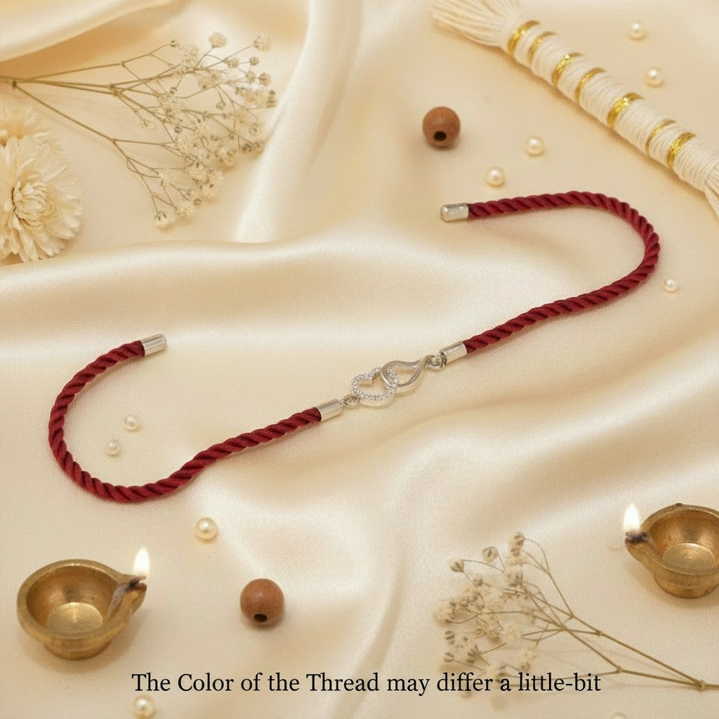 SILVAS sterling silver heart Rakhi with red braided thread on a cream satin background with decorative diyas